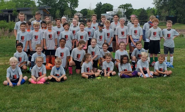 A big group of campers in grey Mo's Soccer Camps shirts on the grass, Summer Camp 2021